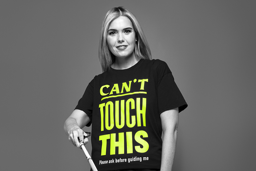 Image of a visually impaired woman called Claire, holding a cane. Claire is wearing a t-shirt that says ‘Can’t touch this - ask before guiding me.’